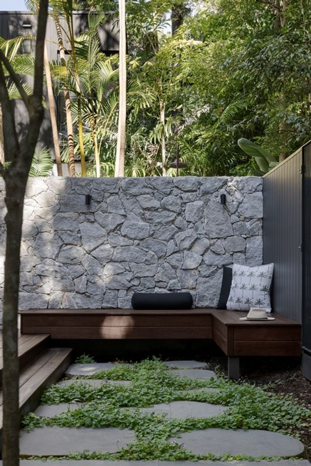 A secluded landscaped courtyard with a built-in dark timber bench wrapping around a corner, set against a rough grey natural-stone wall. Large stepping slabs are softened by low green groundcover, while dense tropical planting and a dark vertical-panel screen create a lush, private outdoor retreat. Small wall-mounted lights and patterned cushions add functional and decorative detail.