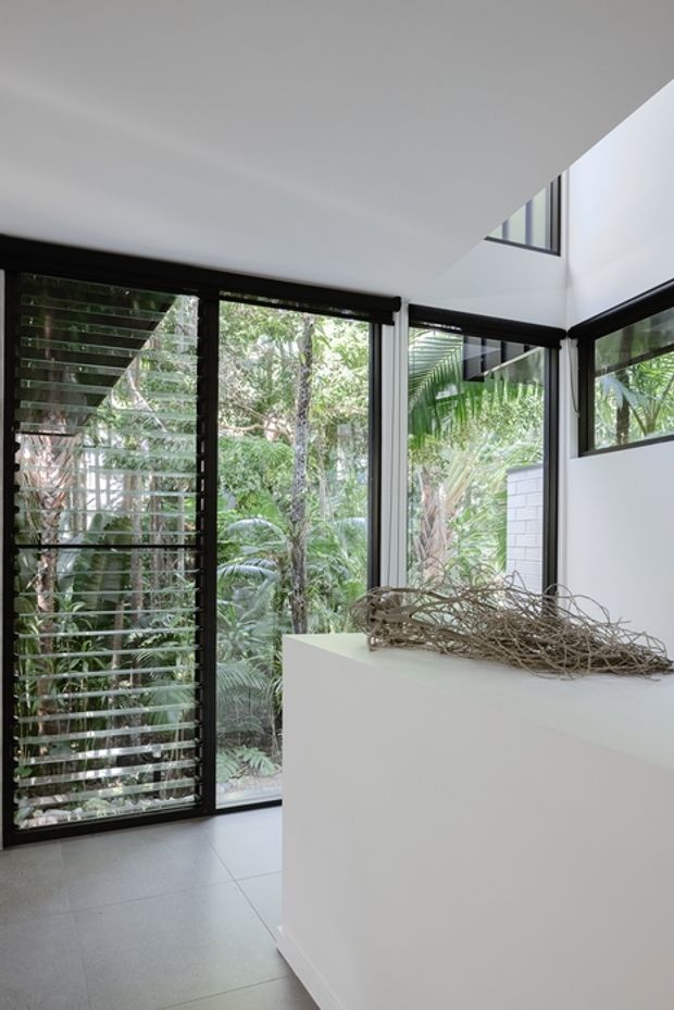A bright, minimalist interior corner with floor-to-ceiling black-framed glazing and horizontal louvres overlooking dense tropical greenery. White walls and a clean white built-in partition contrast with the dark window frames, while a sculptural branch arrangement rests on top. Large pale tiled flooring and a high angled ceiling create an airy, contemporary feel.