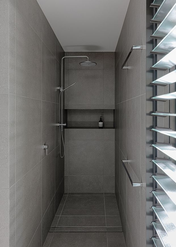 A compact, narrow walk-in shower with dark grey tiled walls and floor, a recessed shelf with a soap bottle, a ceiling-mounted rain shower, and a handheld shower rail. Horizontal louvres on the right provide filtered daylight and ventilation, while the minimalist fittings and continuous tile surfaces create a sleek contemporary bathroom interior.