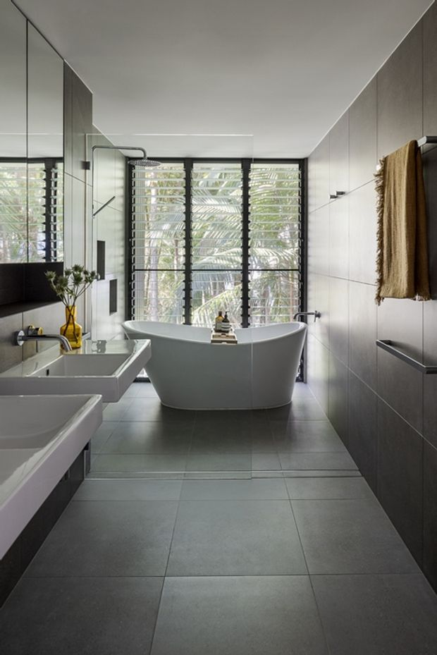 A spacious contemporary bathroom with a freestanding white bathtub positioned in front of full-height black-framed louvred glazing. Two integrated white basins sit along the left wall beneath a large mirror, while dark grey tiled flooring and wall finishes create a refined, monochromatic scheme. A wall-mounted towel and small decorative accents add warmth.