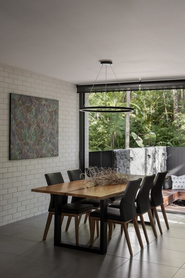 A contemporary dining area with a large timber dining table, black upholstered dining chairs, and a circular pendant light. The room features white painted brick walls, large black-framed glazing opening to lush greenery, a tiled floor, and an adjacent built-in bench seat. A large abstract artwork adds colour to the space.