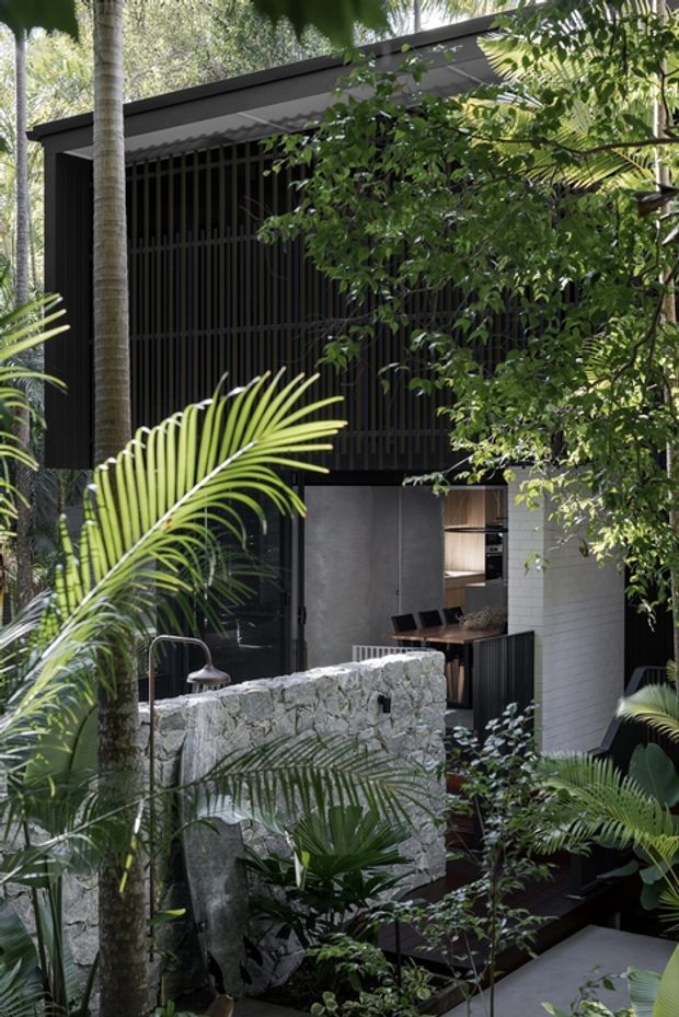 A lush tropical courtyard surrounding a dark, contemporary pavilion or home extension. The building features black vertical screening, a low-pitched roof, dark cladding, and an open outdoor kitchen or entertaining area. A rough natural-stone wall, timber decking, concrete surfaces, and dense palms and foliage create a secluded indoor–outdoor setting.