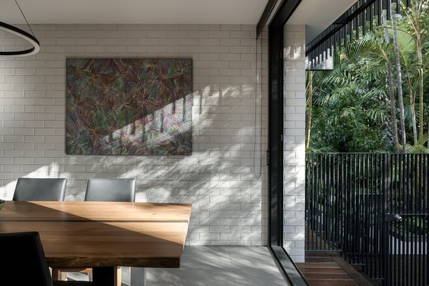 A bright contemporary dining area with a substantial timber dining table and upholstered chairs beside full-height black-framed glazing. The room features a white brick wall with a large abstract artwork, polished concrete flooring, a linear pendant light, and a lush planted exterior visible through the glazing and railing.