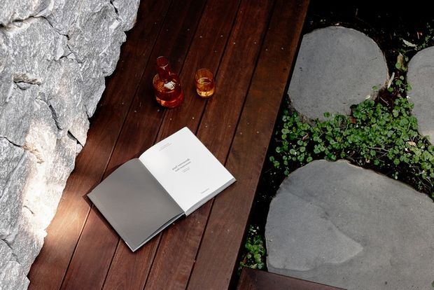 A close-up view of a dark timber deck or boardwalk beside a rugged natural stone wall, with large irregular stepping stones and low groundcover planting alongside. An open book and two small amber drinks rest on the deck, creating a quiet outdoor relaxation setting.
