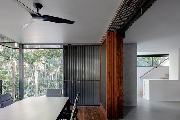 Contemporary open-plan interior featuring a dining area with a large light-coloured table, black chairs, full-height glazing overlooking greenery, a black vertical slatted feature wall, and a warm timber partition. A stairway is visible beyond the opening, with polished grey flooring and a minimalist ceiling fan.