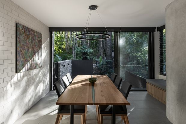 Contemporary dining area centred on a substantial timber dining table with black chairs and a linear pendant light. The room has white painted brick walls, large black-framed glazed doors opening toward dense greenery, a pale tiled floor, and a curved plastered column with built-in timber cabinetry. Natural light and the surrounding vegetation create a calm, refined connection to the outdoors.