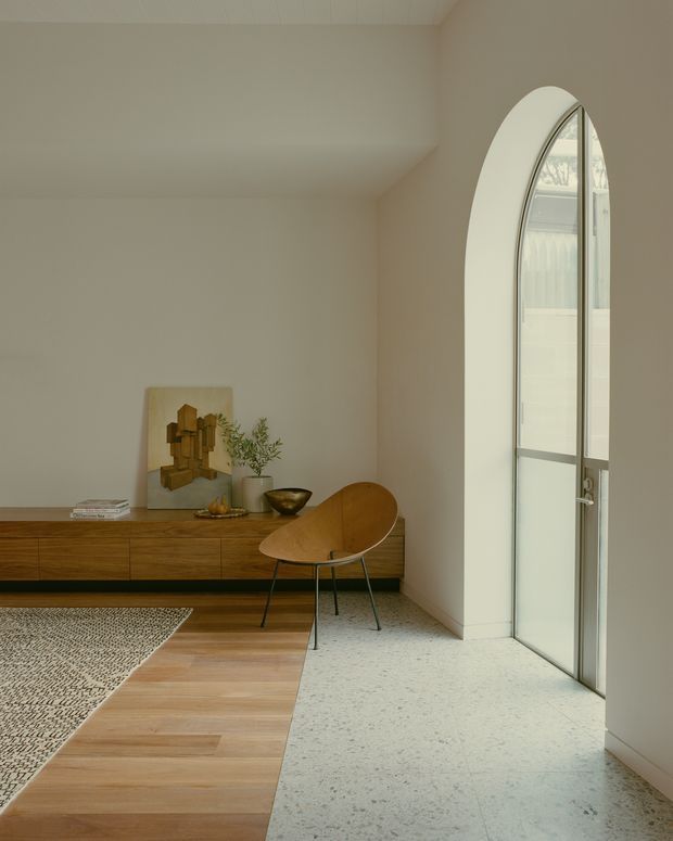 A softly lit minimalist interior corner with a long low timber sideboard, sculptural wooden lounge chair, artwork and potted greenery. A large arched glazed door or window brings in natural light. The floor combines warm timber boards with a pale speckled hard-surface finish, and the palette is calm and neutral.