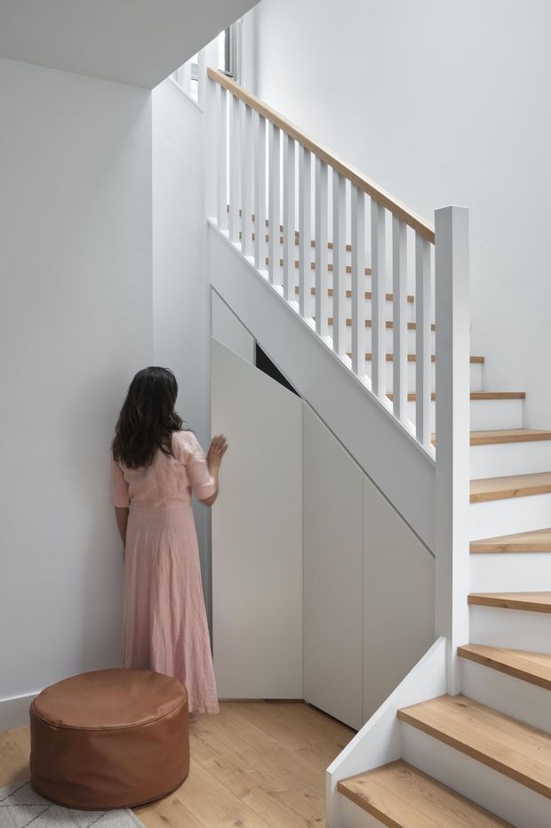 Bright minimalist stairway interior with white walls, a timber-tread staircase, white vertical balustrades, and light built-in storage beneath the stairs. A woman in a pale pink dress stands beside the concealed storage, with a round brown upholstered ottoman in the foreground.