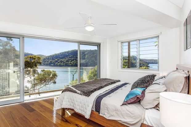 Bright bedroom with a bed positioned beside expansive glazing, overlooking a calm wooded waterfront. The room has polished timber flooring, white walls and ceiling, a ceiling fan, louvre-style windows, and layered neutral bedding with blue accents.