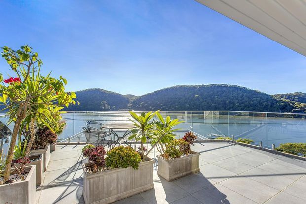 A bright waterfront terrace overlooking a calm body of water and wooded hills. The spacious tiled outdoor area features large concrete planters with tropical and flowering plants, outdoor dining furniture, glass balustrades, and a broad roof overhang.