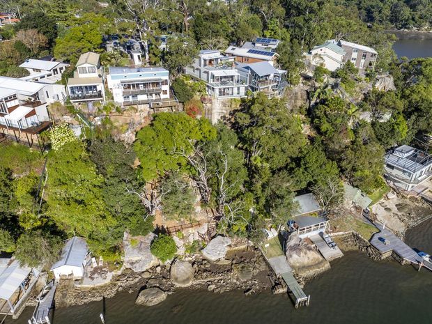 Aerial view of a densely vegetated waterfront residential area, showing several detached homes built along a rocky shoreline with private jetties, waterfront decks and steep, elevated sites. Solar panels are visible on multiple roofs, with the water occupying the foreground.