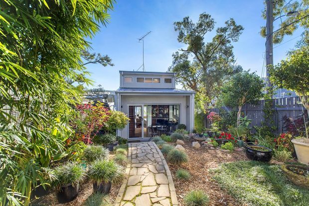 A bright landscaped backyard courtyard leading to a compact two-storey contemporary dwelling. A pale rendered facade has a shallow metal roof, upper clerestory-style windows, and wide glazed sliding doors opening to an interior living space. The garden features a natural stone-paved path, dense planting, ornamental grasses, raised and freestanding planters, and timber fencing.