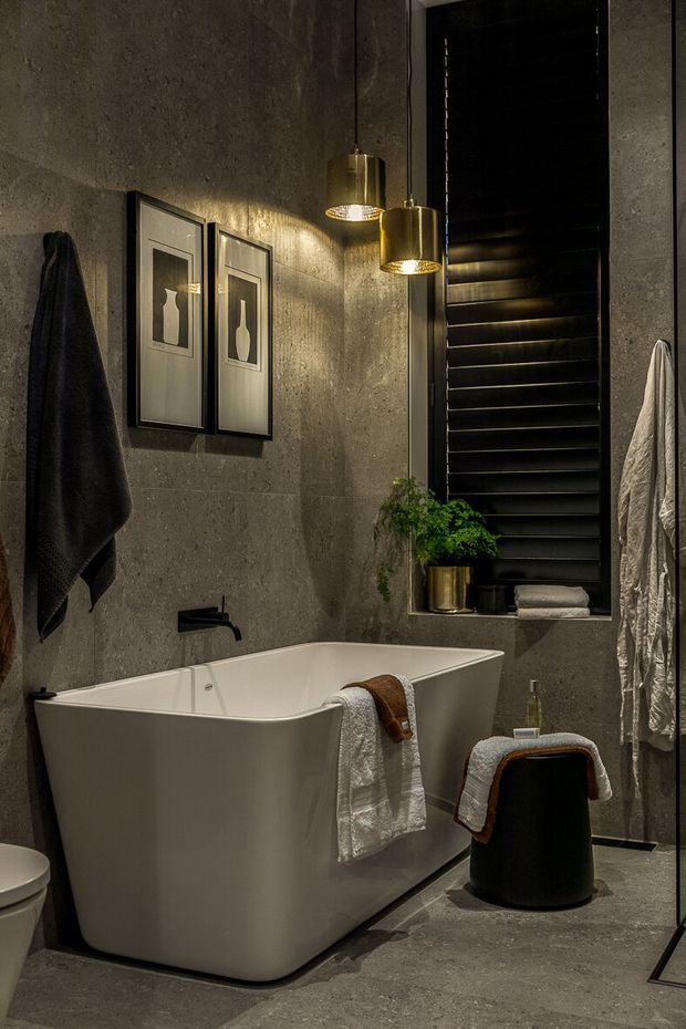 A moody, refined bathroom featuring a freestanding white bathtub, dark grey stone or concrete-look wall and floor finishes, black tapware, a black louvered window covering, and warm brass pendant lights. Framed artwork, towels, a small plant, and a dark stool add detail to the compact space.