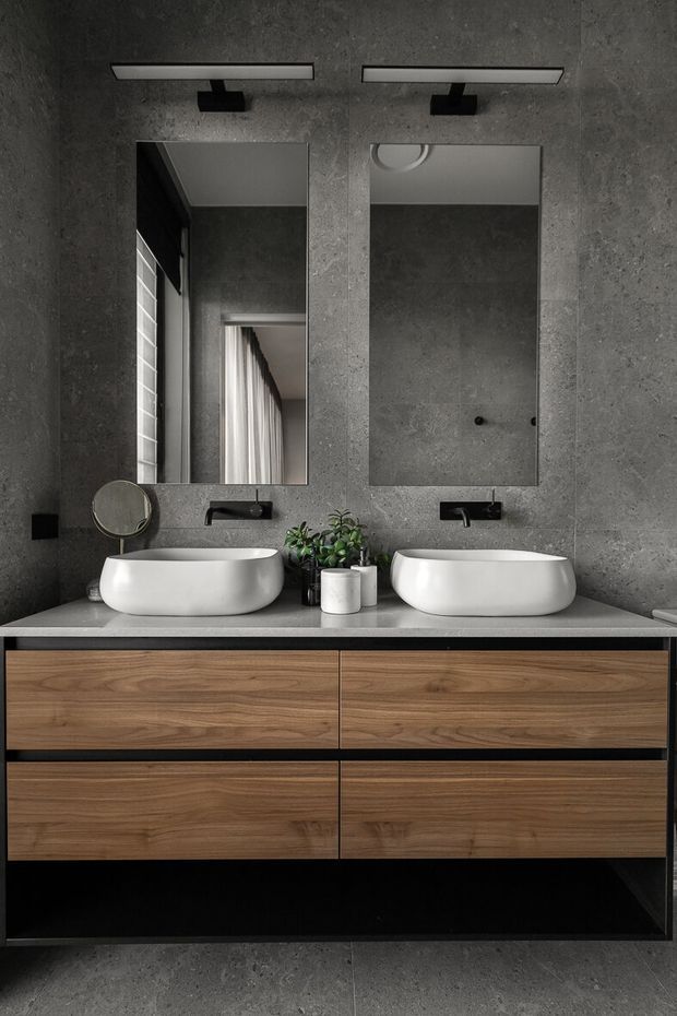 A contemporary double-vanity bathroom with two white vessel basins, matte black wall-mounted taps, rectangular mirrors with integrated horizontal lighting, a grey concrete-look wall and floor, and a floating timber vanity with generous drawers. Small plants and a round magnifying mirror add minor decorative detail.