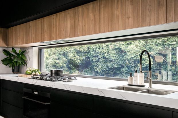 Contemporary kitchen with matte black lower cabinetry, warm timber overhead cabinetry, a white veined stone benchtop, integrated gas cooktop, and a large horizontal window overlooking greenery. A stainless-steel sink with a tall arched tap sits beneath the window, while a potted plant adds a natural accent.