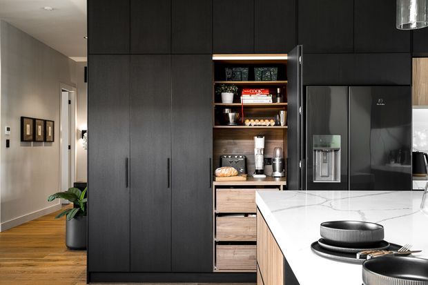 Contemporary kitchen with a large matte black built-in cabinetry wall, integrated open shelving and appliances, and a white veined stone island. Warm timber flooring and timber drawer fronts add contrast, while a hallway is visible beyond the kitchen.