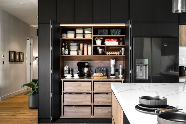 A contemporary kitchen focused on a tall, dark built-in pantry and appliance storage wall. The open pantry features timber shelving, drawers, countertop appliances and food storage, with a large dark refrigerator and a white stone island in the foreground. Warm timber flooring and neutral walls balance the dark cabinetry.