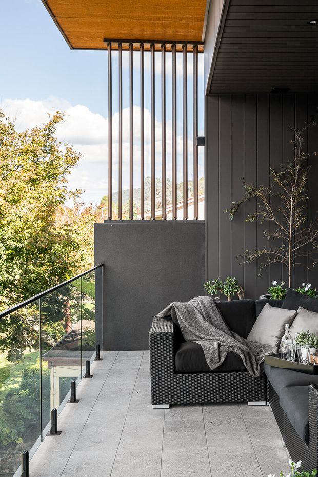 A contemporary covered outdoor balcony or terrace overlooking leafy surroundings. The space features large-format grey tiled flooring, a dark vertical-panel exterior wall, a timber-lined soffit, slender vertical metal balustrade elements, and clear glass balustrading. Dark woven outdoor lounge furniture with grey cushions and a throw creates a comfortable seating area, with potted plants adding greenery.
