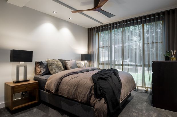 A contemporary bedroom with a large upholstered bed, layered neutral bedding, bedside tables and lamps, a wall-mounted television, dark carpet, and floor-to-ceiling sheer curtains opening to a leafy exterior. Recessed ceiling lights and a ceiling fan add practical illumination and ventilation, while the restrained palette creates a refined, calm atmosphere.