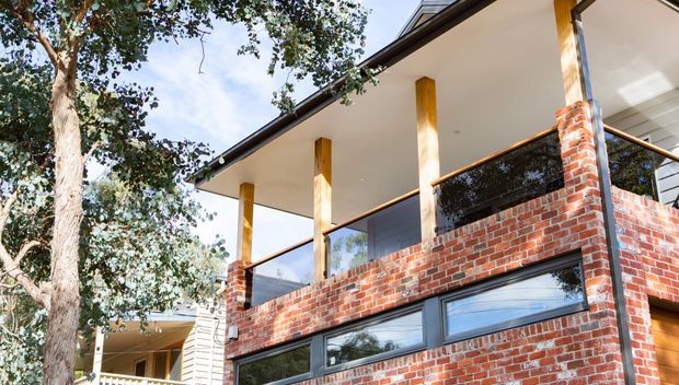 Exterior architectural view of a contemporary two-storey brick residence, featuring a cantilevered upper-level balcony with dark-framed glazing and balustrade, slender timber-toned columns, broad roof overhangs, and horizontal lower-level windows. Mature trees partially frame the building.