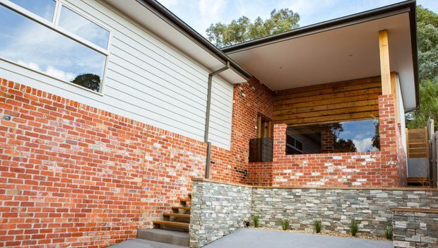Exterior view of a contemporary residential entry or covered outdoor area. The facade combines red brickwork, white horizontal weatherboard cladding, large windows, timber detailing, and a low-pitched roof with a broad sheltered overhang. Stone-clad retaining walls and steps lead up to the recessed opening.