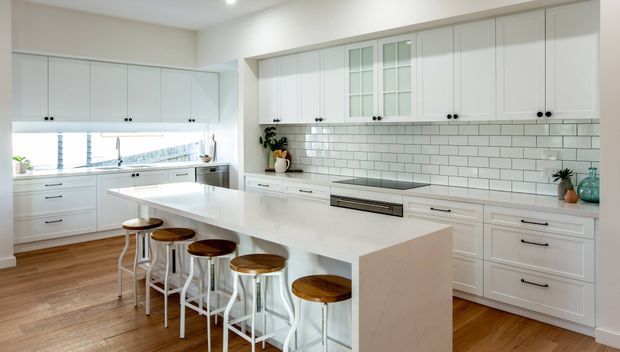 Bright, spacious white kitchen with extensive shaker-style cabinetry, glass-front upper cabinets, a subway-tile backsplash, stone island bench, integrated appliances and timber flooring. The large island includes seating for five or more on simple stools, with abundant natural light and a clean, classic coastal feel.