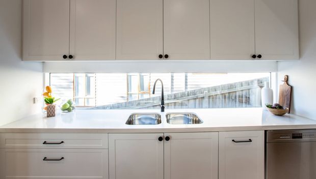Bright contemporary kitchen with white flat-panel cabinetry, a long white benchtop, double undermount sink, dark tapware, and integrated upper storage. A wide horizontal window provides a view toward adjacent buildings, with minimal countertop styling and a stainless-steel dishwasher at right.