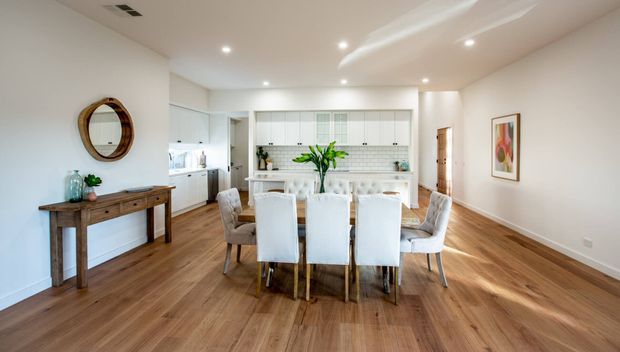 Bright open-plan dining area with a six-seat dining table and upholstered chairs, connected directly to a modern white kitchen. The room features warm timber flooring, white walls and cabinetry, recessed ceiling lights, a timber console, a round mirror and framed artwork.