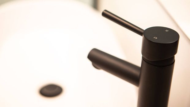 Close-up product-style image of a matte black cylindrical bathroom tap mixer with a slim lever and side spout, positioned over a white ceramic basin. A dark circular basin drain is visible in the background.