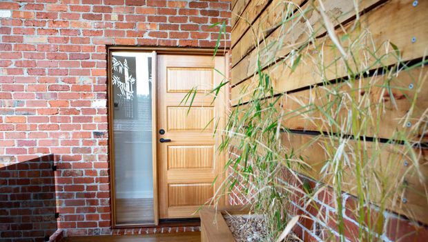 Close view of a residential entryway with a timber-panelled front door and narrow glazed sidelight, set into a red brick wall. A horizontal timber-clad wall and planter with tall grasses frame the entrance, creating a warm textural exterior threshold.