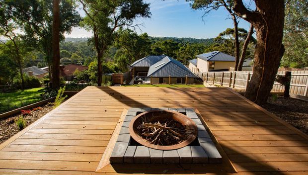 A spacious elevated timber deck surrounded by mature trees, with a central built-in circular fire pit set into a dark paver surround. The deck overlooks a leafy suburban or bushland setting and nearby pitched-roof homes, creating an outdoor entertaining area.