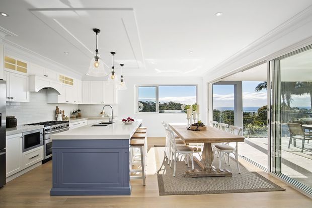 Bright coastal-style open-plan kitchen and dining area with white Shaker cabinetry, a muted blue-grey island, white subway-tile splashback, timber flooring, pendant lights, and a rustic timber dining table. Large windows and sliding glass doors provide abundant natural light and views toward the sea and palms, with an outdoor terrace visible beyond.