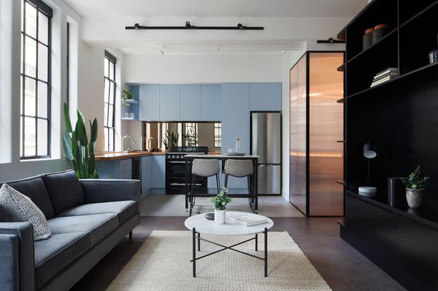 A bright open-plan loft-style interior combining a living room, kitchen and dining area. The space features large black-framed industrial windows, pale blue vertical-panel kitchen cabinetry, dark timber or black built-in shelving, a compact island with stools, a dark sofa, and a warm backlit glazed partition. A textured light rug and round coffee table soften the darker flooring and joinery.