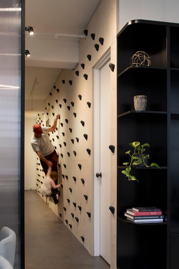 Interior view of a narrow hallway featuring a full-height indoor climbing wall with black climbing holds, used by an adult and child. The wall is clad in pale timber panels with visible fixings and integrated climbing access around a white door. A tall black open shelving unit displays books, a plant, decorative objects, and storage. The space has a polished grey concrete floor, white ceiling, track-style spotlights, and a contemporary functional design.
