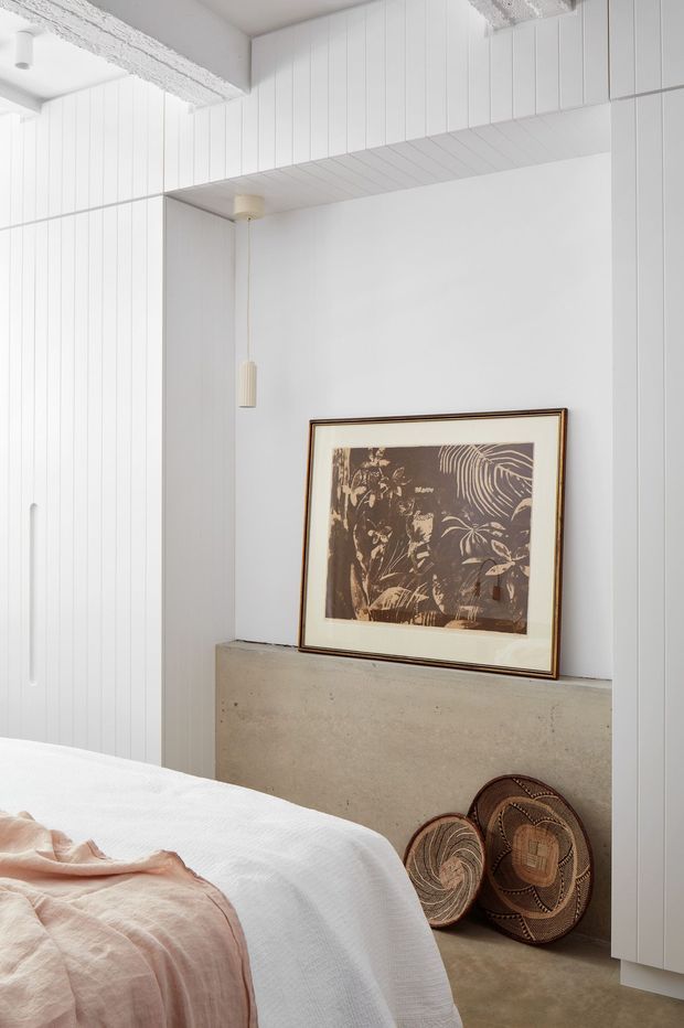 A bright, minimalist bedroom corner with white vertical wall panelling and a white overhead soffit creating a recessed niche. A raw concrete ledge supports a framed botanical artwork, while woven baskets rest below. The foreground shows part of a bed with white bedding and a soft blush-pink throw. Slim pendant lights add subtle warmth to the otherwise neutral interior.