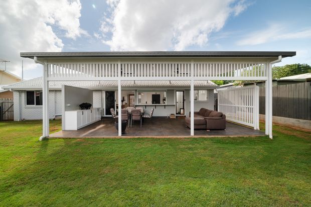 Exterior view of a single-storey home with a large covered outdoor entertaining area. The white pavilion-style structure has a flat roof, vertical slatted screening, white posts and railings, an outdoor kitchen or bar, dining furniture, and lounge seating opening onto a lawn. The existing house is visible behind it, with neighbouring fencing and greenery in the background.
