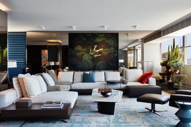A spacious, high-end contemporary living room with a large L-shaped sectional sofa, sculptural coffee tables, a patterned area rug, accent chairs, floor-to-ceiling glazing, and a large botanical artwork feature wall. The room has a bright neutral palette with blue and red accents, integrated linear ventilation, and layered modern lighting.