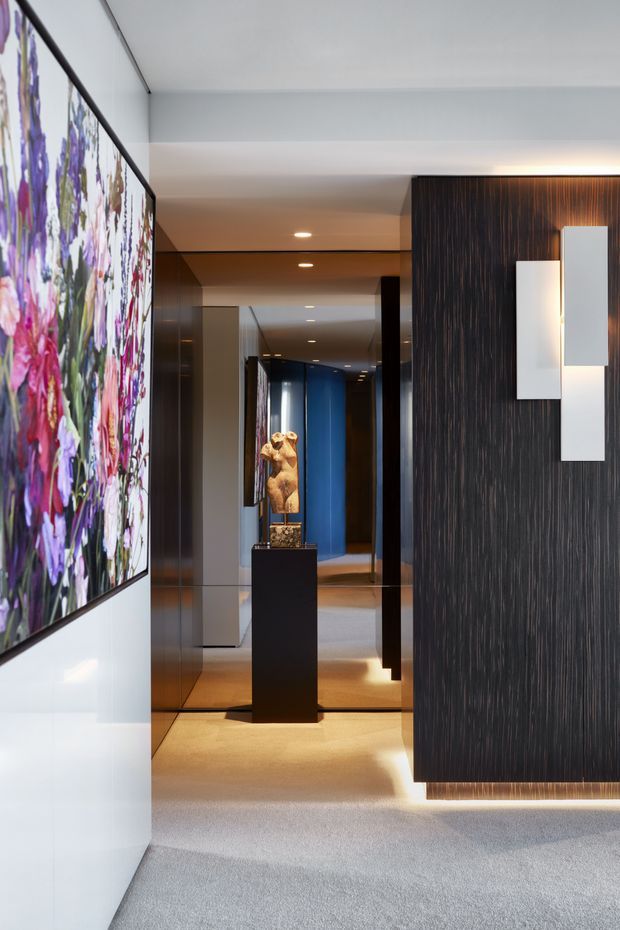 A refined contemporary interior hallway or entry corridor with grey carpet, white walls, mirrored panels, recessed ceiling lights, and dark vertically textured timber wall panelling. A large colourful floral artwork occupies the left wall, while a sculptural object stands on a black pedestal in the corridor beyond. Geometric white wall sconces and concealed linear lighting add a polished, gallery-like atmosphere.