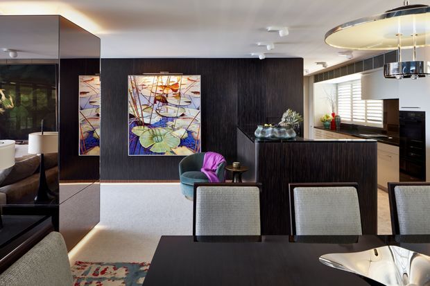 A refined open-plan dining and kitchen interior with dark timber wall panelling and cabinetry, a large dining table with upholstered chairs, colourful abstract artwork, and a sculptural pendant light. The kitchen features a dark island, integrated appliances, white cabinetry, and horizontal window blinds. A lounge area is partially visible through the open space and mirror reflections.