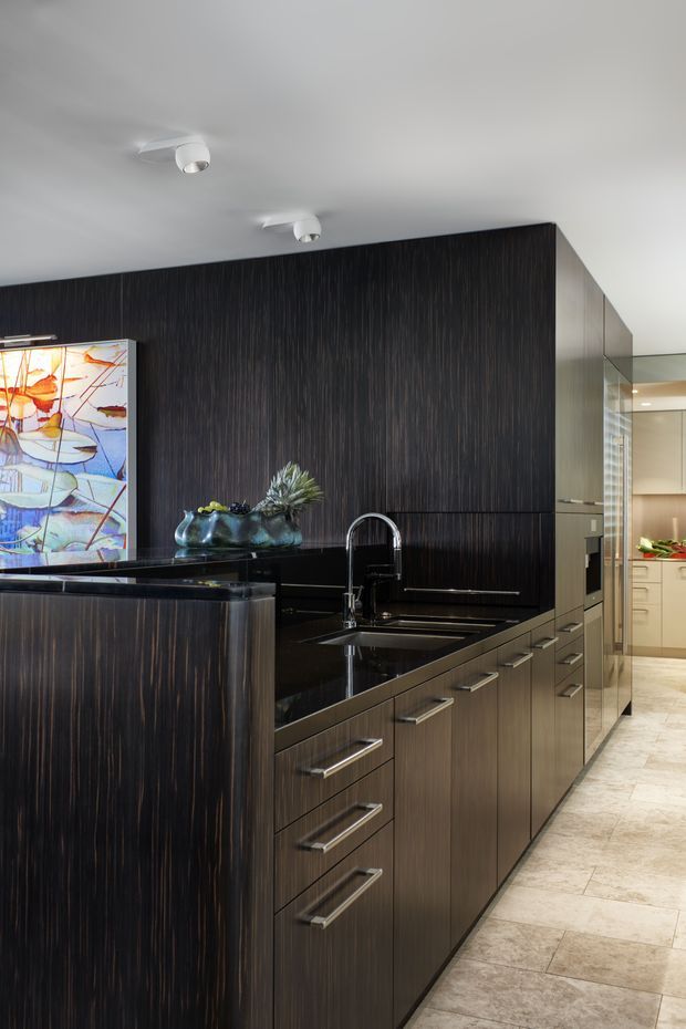 A sophisticated contemporary kitchen with dark vertically textured cabinetry, a glossy black stone island benchtop, integrated sink and tall curved mixer tap. The kitchen includes extensive handle-fronted joinery, a built-in oven wall, pale stone-look flooring, and a colourful abstract artwork at the edge of the frame. A secondary lighter kitchen or pantry area is visible through the opening at right.