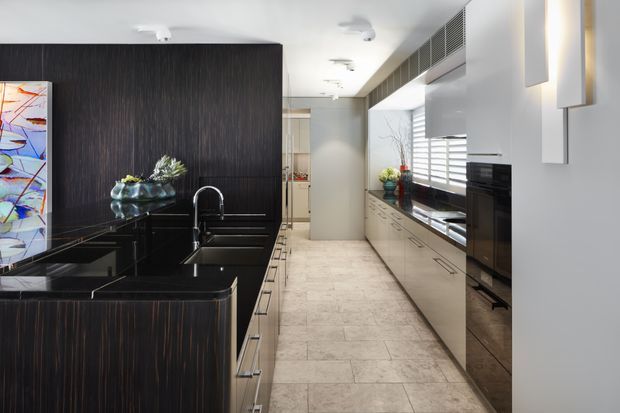 Contemporary galley kitchen with a long black stone island and dark vertically grained cabinetry on the left, paired with light cabinetry and integrated appliances along the right wall. The space features pale stone-look flooring, white walls, horizontal window blinds, linear ventilation grilles, and minimalist built-in wall lighting. A colourful artwork and small decorative objects add contrast.