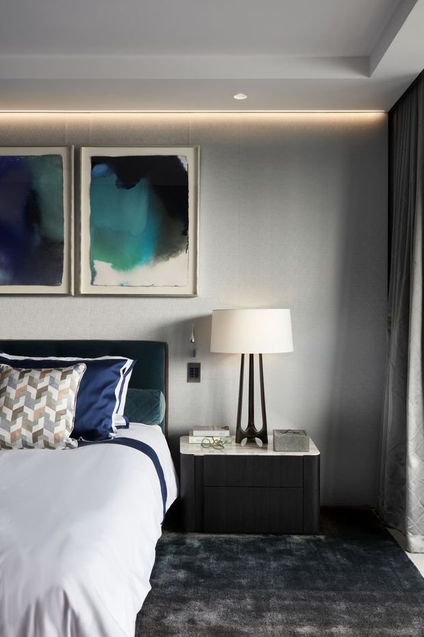 A refined contemporary bedroom vignette featuring a partially visible upholstered bed with crisp white and navy bedding, patterned accent pillows, a dark bedside table with a sculptural tripod lamp, abstract blue-green artwork, textured grey walls, recessed cove lighting, dark carpet, and full-height curtains.