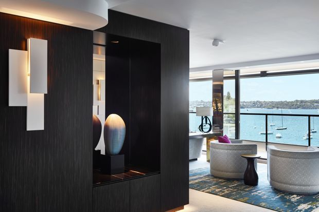 Refined contemporary living room with dark vertical timber feature joinery and integrated display niche, sculptural wall lighting, and two rounded lounge chairs beside expansive waterfront glazing. A blue patterned rug and restrained furnishings create a polished, serene interior overlooking a bay.