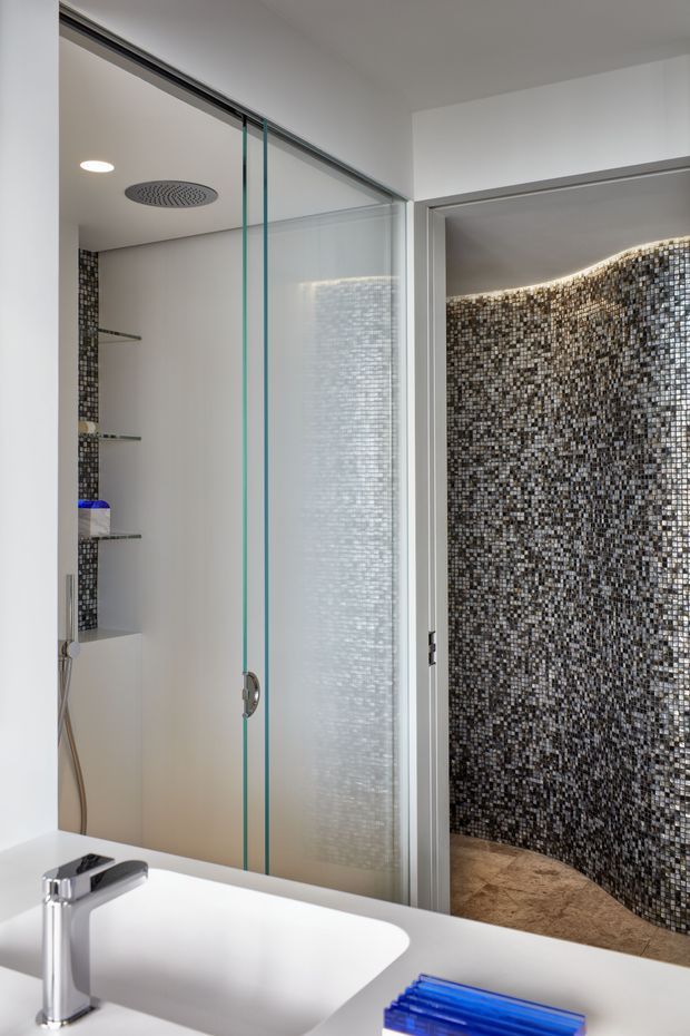 A contemporary bathroom featuring a white integrated basin and chrome tap in the foreground, a frameless glass shower enclosure with a ceiling-mounted rain shower, recessed shelving, and a curved wall finished in small dark mosaic tiles. The palette is predominantly white, grey, black, and chrome with a warm stone-like shower floor.