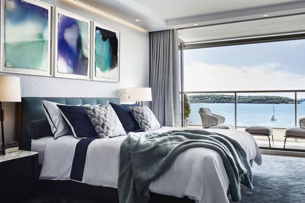 A refined waterfront bedroom with a large upholstered bed facing floor-to-ceiling glazing and a balcony or terrace overlooking calm water. The room features layered white and navy bedding, a textured blue-green throw, abstract blue artwork, bedside lamps, full-height curtains, and a soft grey rug.