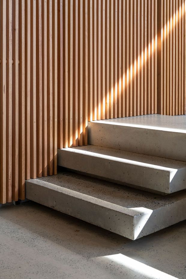 A close-up architectural interior detail showing a minimalist concrete stairway ascending beside a wall clad in evenly spaced vertical timber slats. Strong sunlight filters through the slatted screen, casting rhythmic stripes of light and shadow across the concrete treads and floor. The composition emphasises raw material texture, geometric lines, and the junction between timber screening and cast concrete.