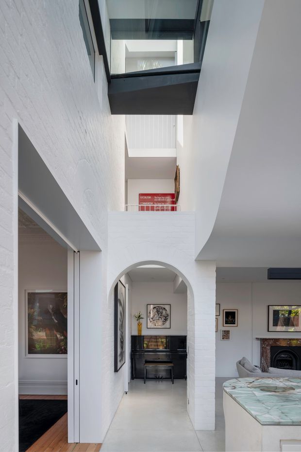 Bright interior view of a multi-level residential hallway and living area, featuring white-painted brick walls, a large arched opening, polished light flooring, black-framed overhead glazing, and a black upright piano beneath a mezzanine. The adjoining living room includes a fireplace, framed artwork, light seating, and a prominent pale stone island or counter in the foreground.
