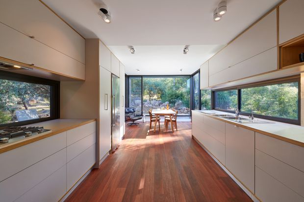 A bright, contemporary galley kitchen with extensive white built-in cabinetry, integrated appliances, timber benchtop accents, and long horizontal windows. The kitchen opens toward a dining area with a timber table and chairs, while large glazed openings provide views to surrounding trees. Warm timber flooring and discreet recessed and track lighting create a refined, natural interior.
