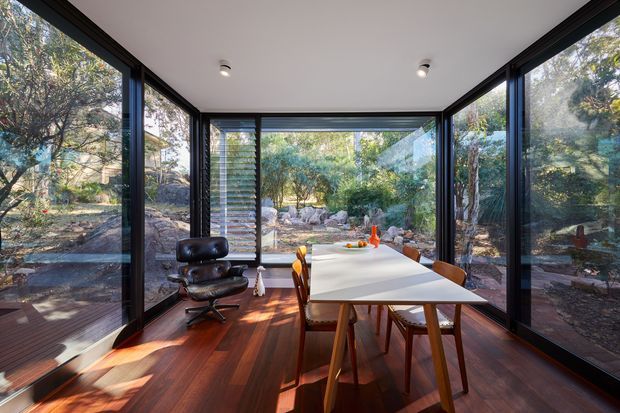 A bright, glass-enclosed dining room or sunroom surrounded by landscaped bush, with full-height black-framed glazing on three sides, a white rectangular dining table, timber dining chairs, a black leather lounge chair, and rich dark timber flooring. The space has a clean contemporary architectural character and strong indoor-outdoor connection.