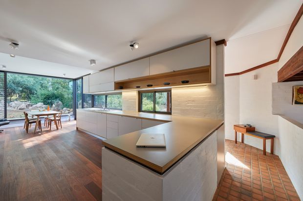 Open-plan kitchen and dining area with a large central island, integrated pale cabinetry, timber detailing, and a warm textured wall finish. The kitchen opens to a glazed dining space overlooking leafy landscaping, while a terracotta-tiled adjoining area with built-in shelving is visible on the right.